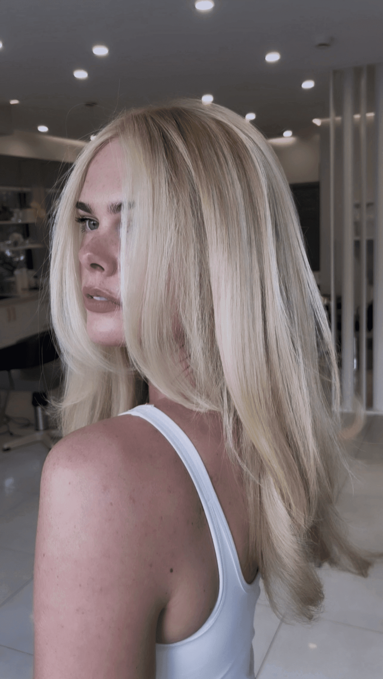 Blonde woman with long hair in a modern salon, wearing a white tank top.