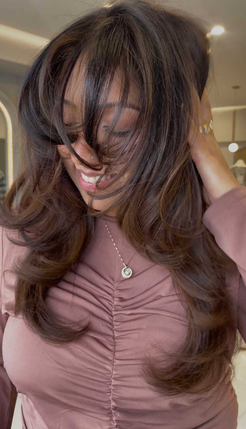 Woman smiling with long, wavy hair and wearing a ruched pink blouse.