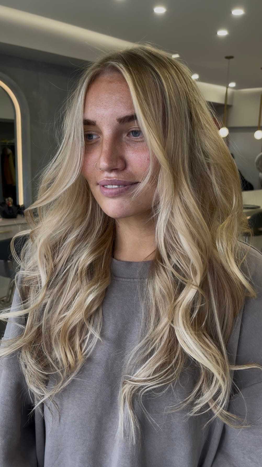 Blonde woman in a salon with long, wavy hair wearing a gray sweatshirt.